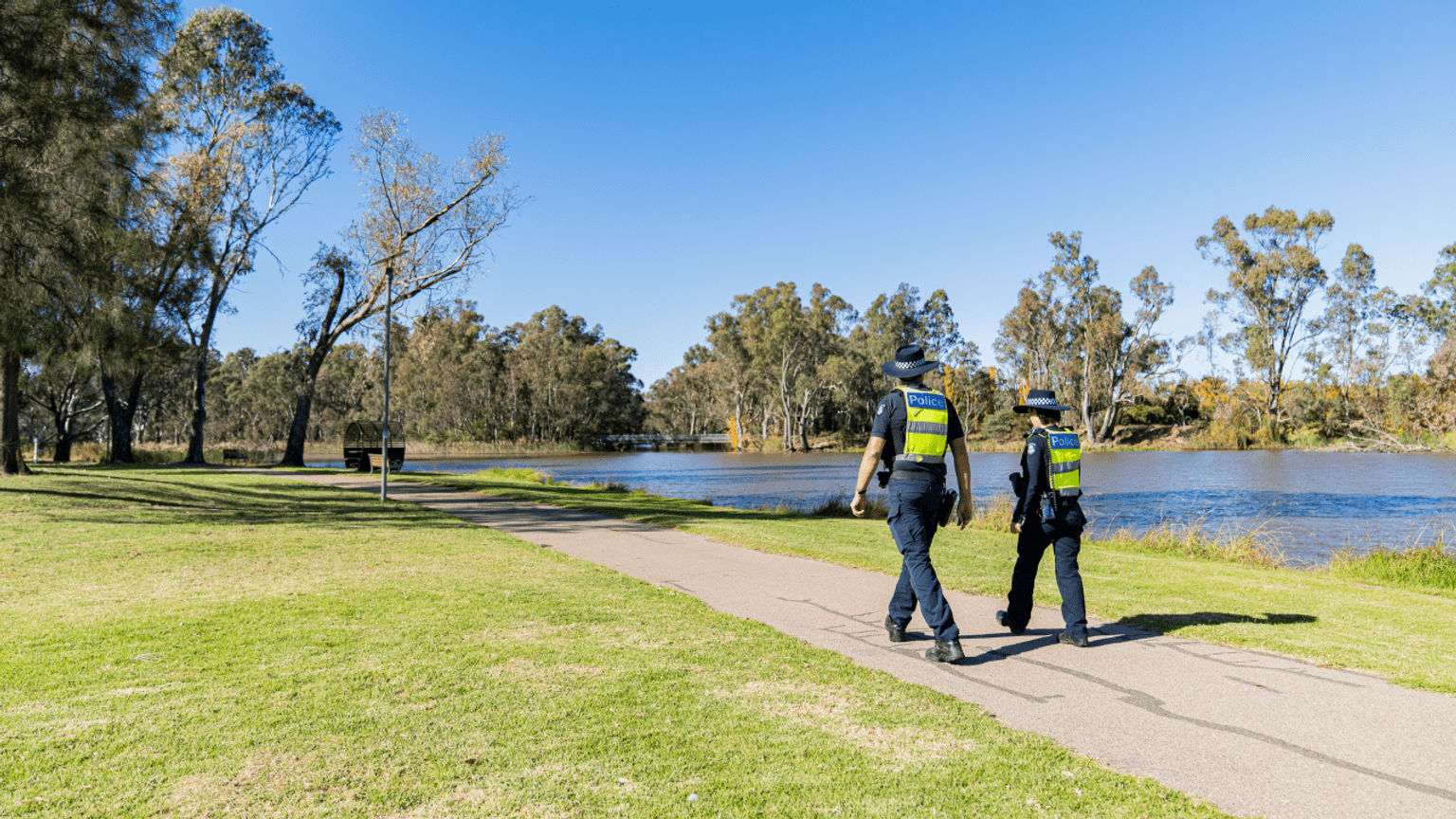 Two police officers in police uniform, walking in the sun on a path next to the river.