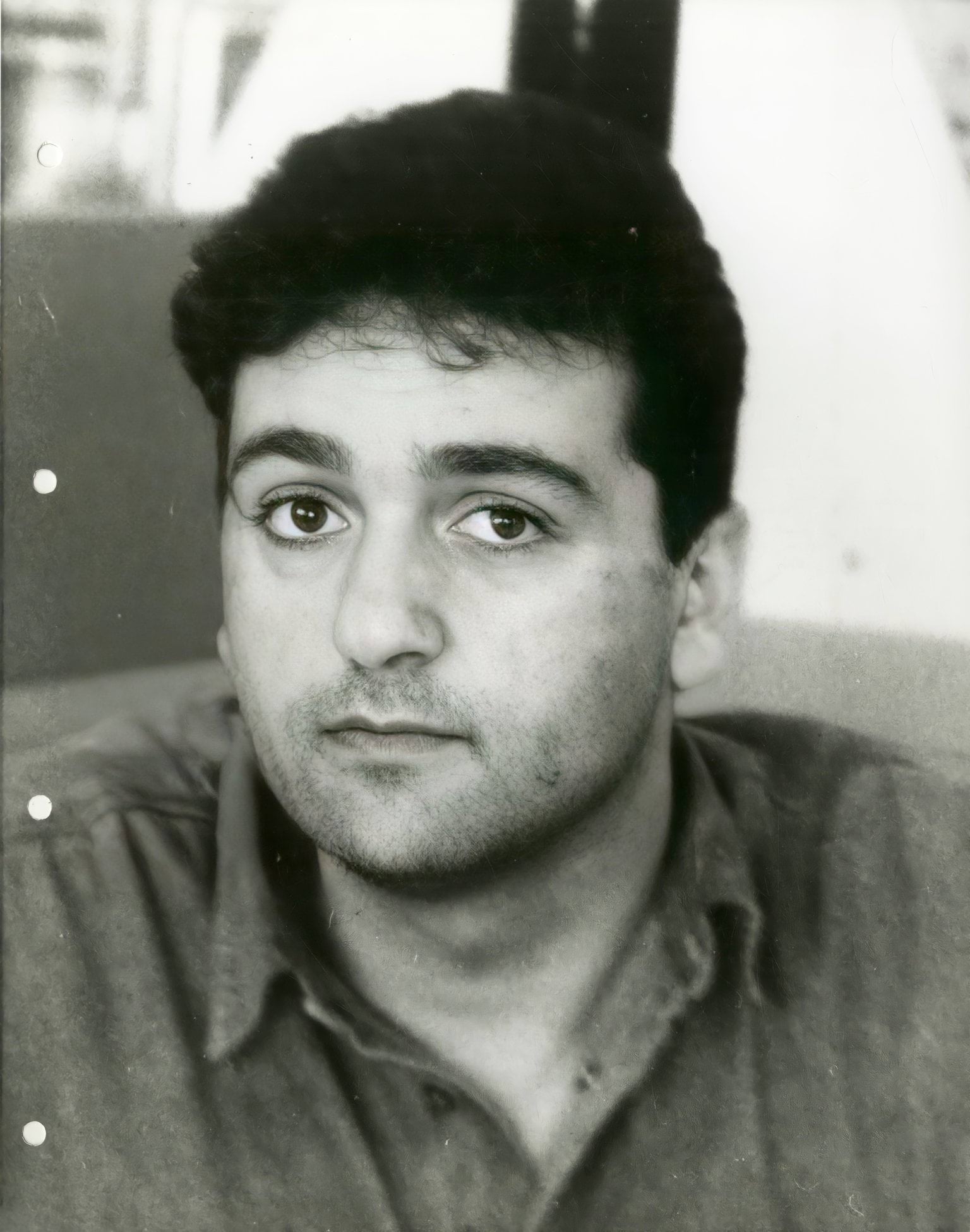 A black and white photo of 26-year-old white man looking directly at the camera. He has short dark curly hair, has slight stubble and dark eyes. The photo looks to be from the early 1990's.