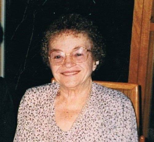 An older photo showing a older woman with very short and curly darker hair sits in a chair, smiling directly at the camera. Her hands are clasped in her lap, and she is wearing glasses, a watch and floral (or dotted) top.