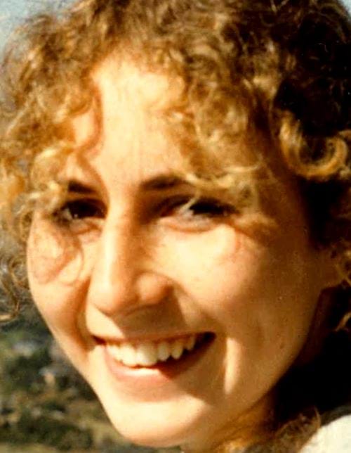 A 23-year-old white woman with curly dark blonde hair is turning and smiling at the camera. She has brown eyes and is fresh faced.