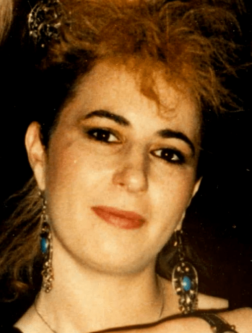 A 23-year-old white woman with dark eyes and eyebrows is looking directly at the camera. Her hair is feathered and looks dark on the side and blonde at the top and the back. She is half smiling, and wearing 1980's dark eye makeup and peach lipstick, along with ornate gold dangling earrings with blue stones.