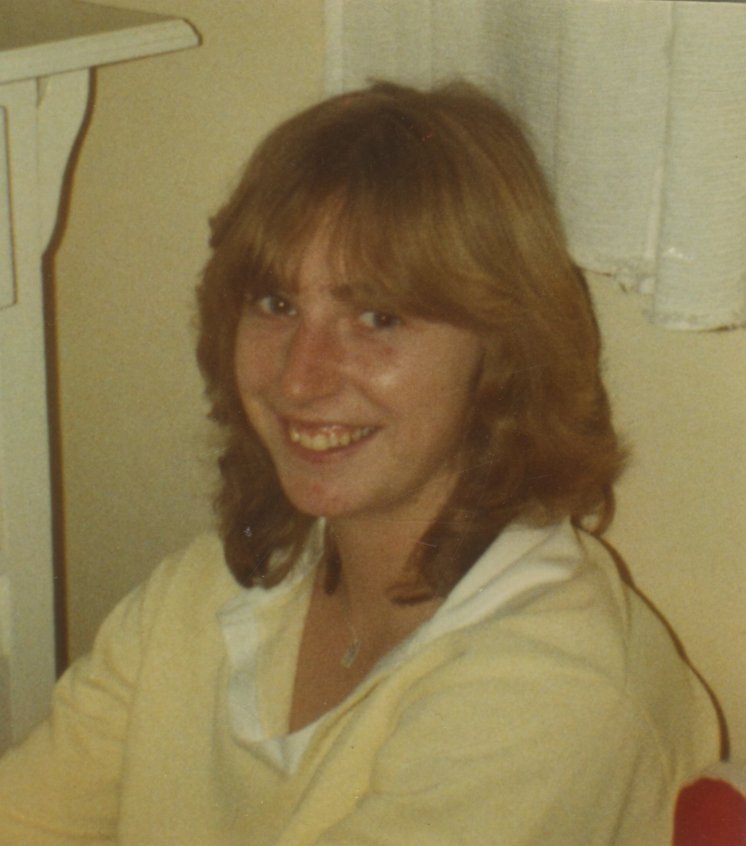 A 25 year old white woman with feathered dark blonde hair is sitting and smiling at the camera. She is wearing a pale yellow polo shirt with a white collar.