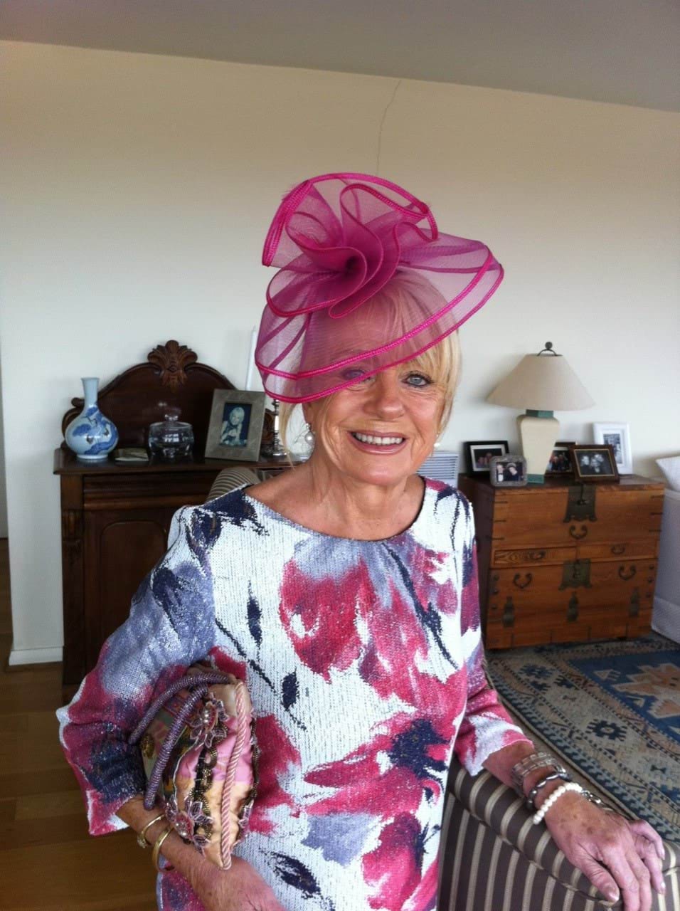 A 69-year-old Caucasian woman with blonde hair is looking directly at the camera and smiling with teeth. She is wearing a pink fascinator which obscures her right eye, and a white 3/4 quarter length sleeved white dress with pink and purple flowers on it. She has blue eyes and is wearing black eyeliner.