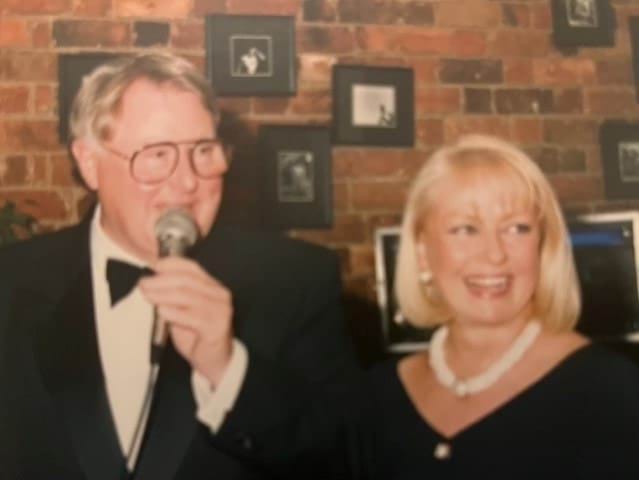 A 69-year-old Caucasian woman is looking away from the camera and smiling. She has blonde, shoulder-length hair and blue eyes. She is wearing a white necklace and a black top. She is standing next to a Caucasian man of approximately the same age wearing a tux, bowtie and glasses. He is holding a microphone and looking away from the camera.