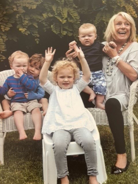 A 69-year-old Caucasian woman is looking away from the camera and smiling widely. She has blonde, shoulder-length hair with a side fringe, and blue eyes. She is wearing a white beaded necklace, a white top, black leggings and black ballet flats. she is holding a baby in her hands and is seated next to three other toddlers on white cane outdoor furniture, on a green lawn.