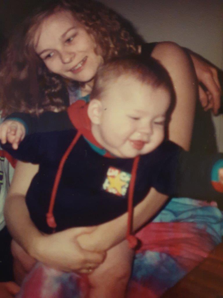 Shari Davison, a 27-year-old white woman is cuddling a toddler in her lap while smiling and looking away from the camera. She has dark blonde curly hair, worn loose, and is wearing a pink and blue dress that looks to be tie-dyed. The photo looks seems to be from the late 1980's or early 1990's.