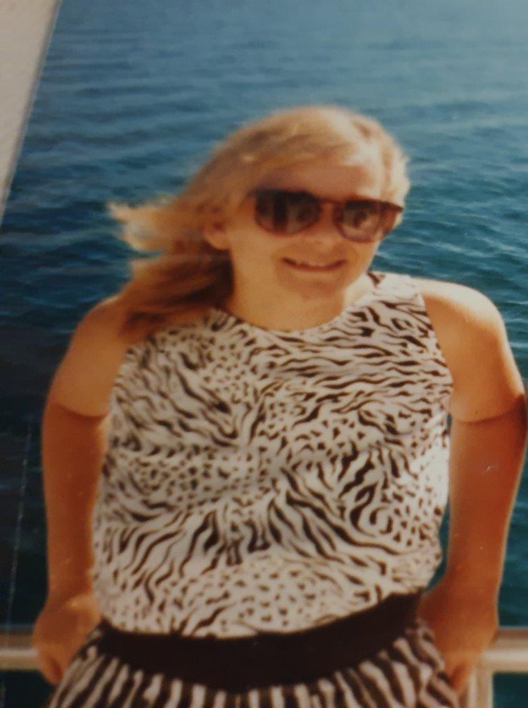 A 27-year-old white woman is wearing dark sunglasses and smiling at the camera. She has blonde layered hair, and is leaning back on a railing in front of water (possibly on a boat). She is wearing a black and white leapoard print sleeveless dress. The photo looks to be from the 1990's.