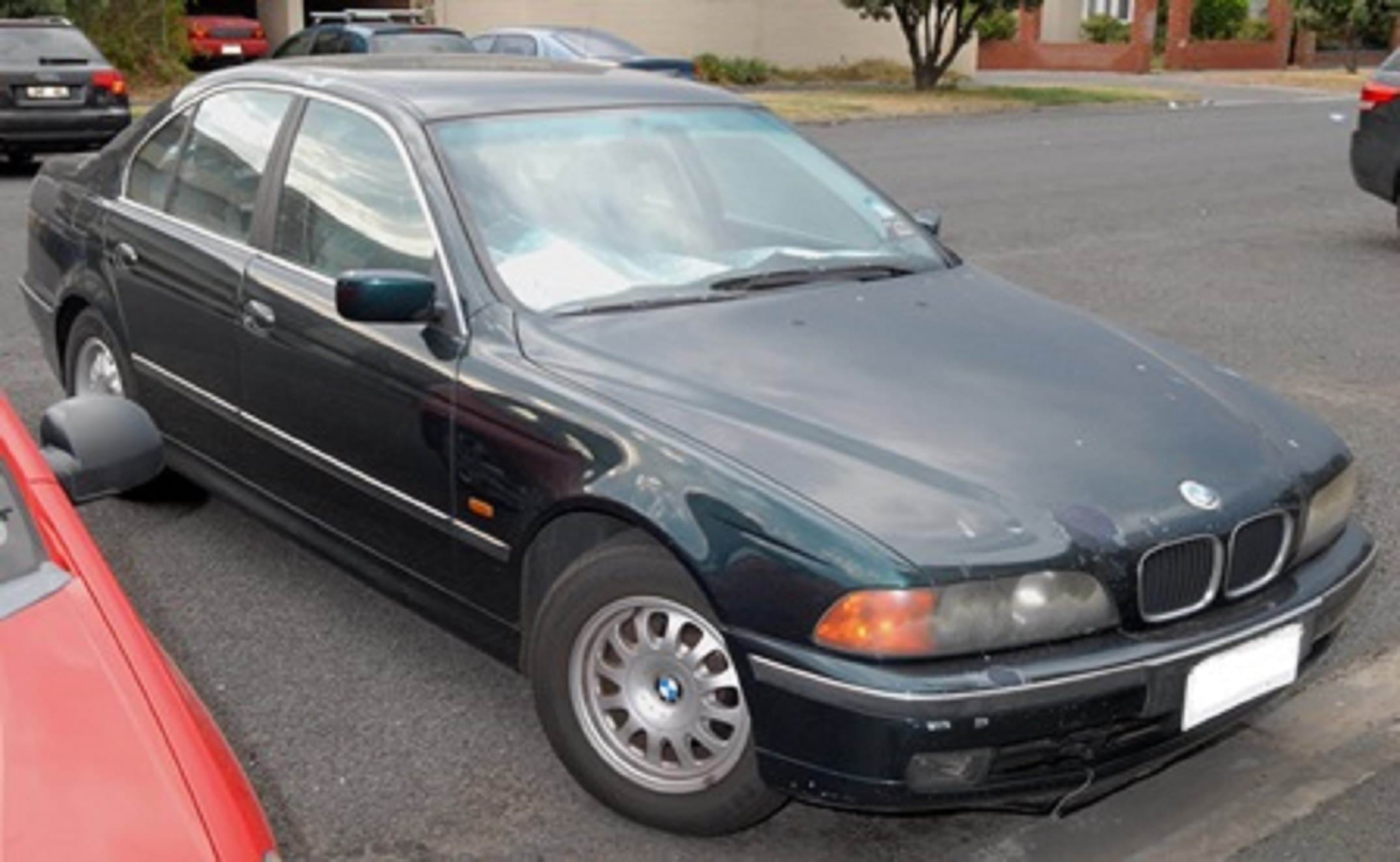 A 1997 dark green BMW is parked on a residential street. There is a silver sun shade on the inside of the windscreen that has partially fallen down. The paint on the car's bonnet is scratched and faded in some spots.