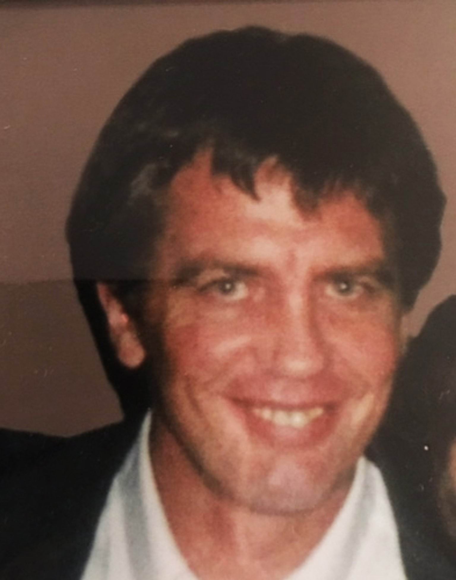 A photo of missing person Selman (Sam) Mala. The photo shows a man with medium length very dark brown or black hair, of Caucasian appearance. He is smiling and looking directly at the camera. He is wearing a white dress shirt and black dress jacket. The photo appears to be from the 1990s. He appears to be in his early or mid 30s.
