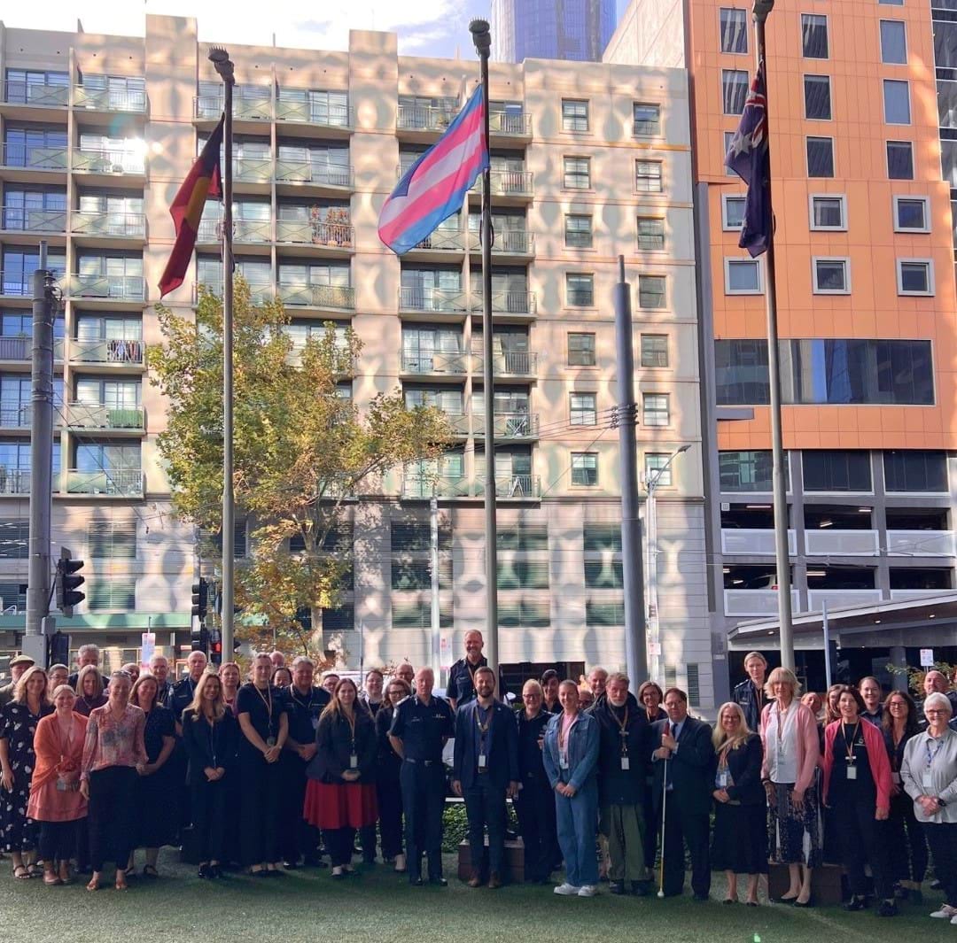 Members of the community stand under the raised Trans flag, Aboriginal flag, and Australian flag. 