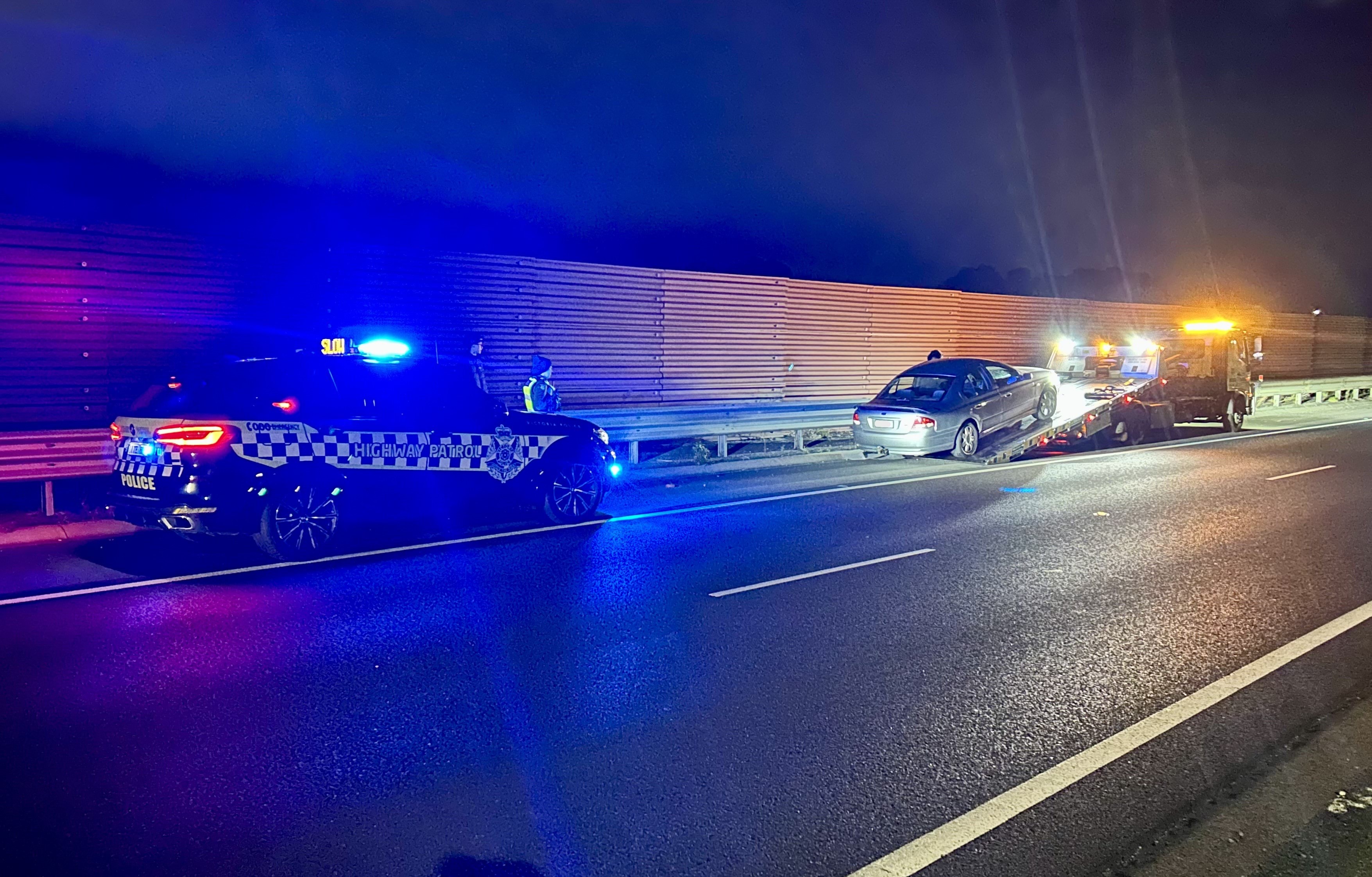 A Ford Falcon gets pulled onto the back of a tow truck. A police car sits behind it with its lights activated.