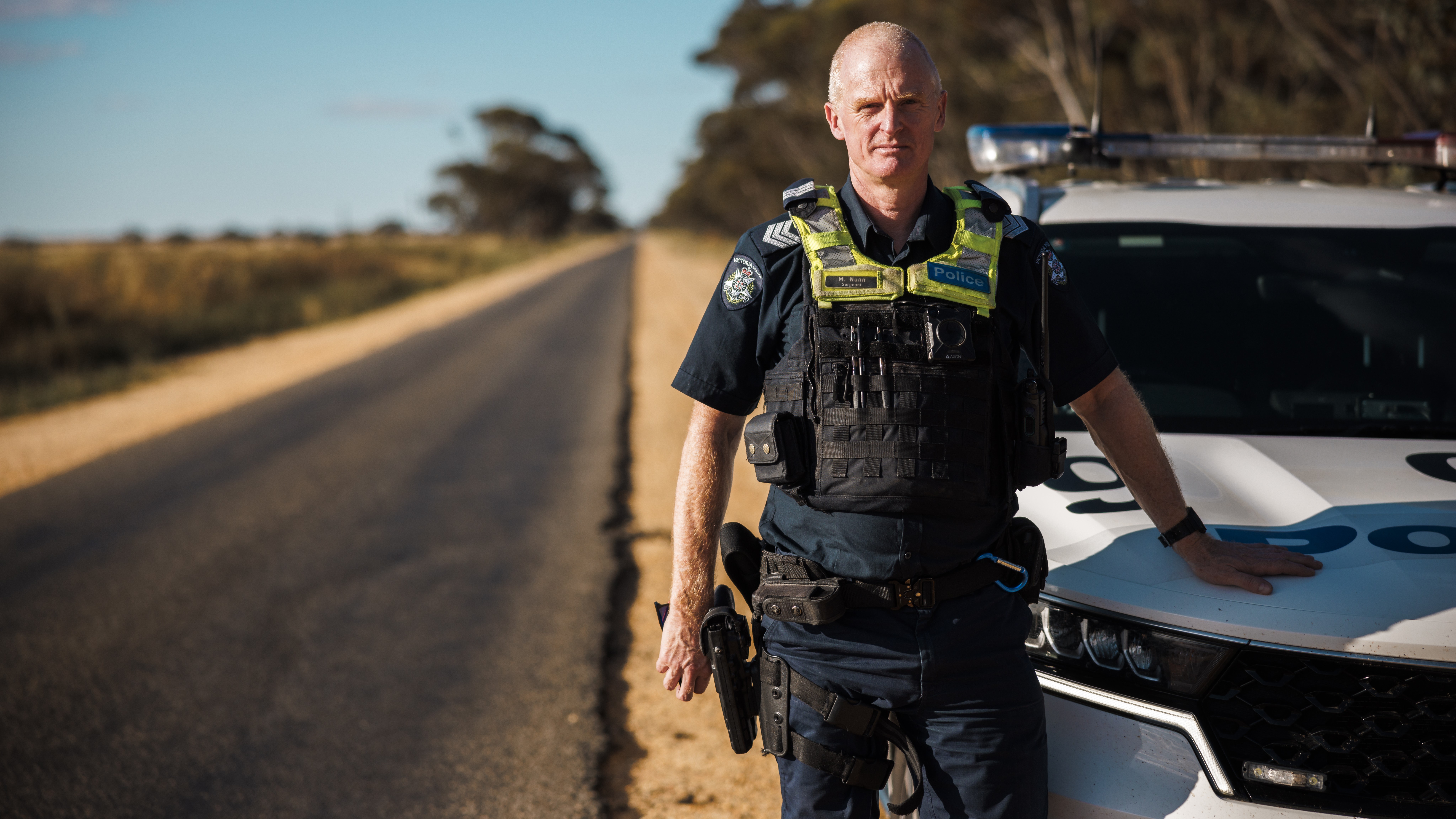 Police Life - image of male police officer fully kitted up standing next to a police car that is parked on the side of a rural road.