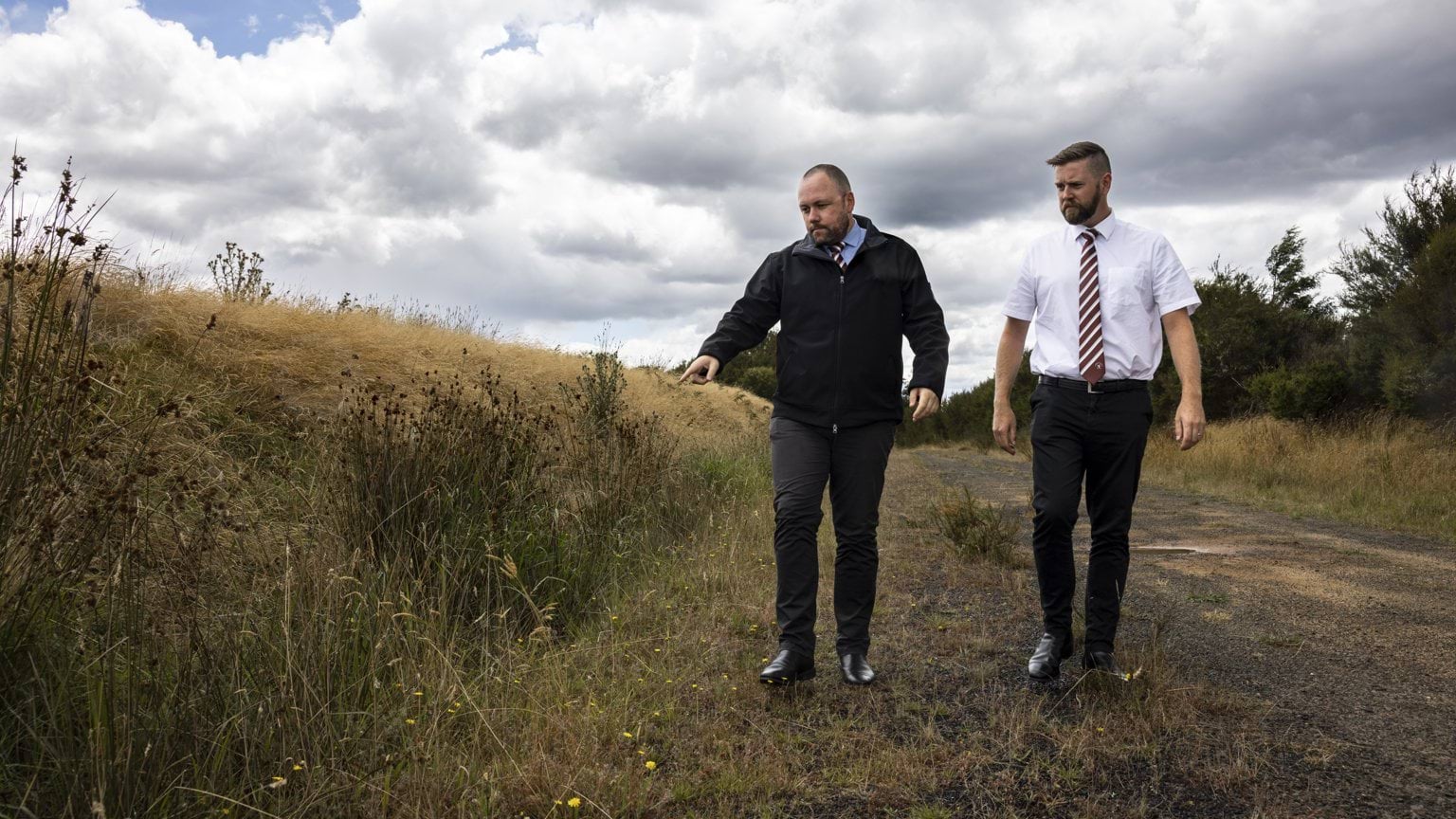 Police Life -- two detectives in plain clothes are walking along a dirt road. One detective is pointing to grass scrub beside the road.