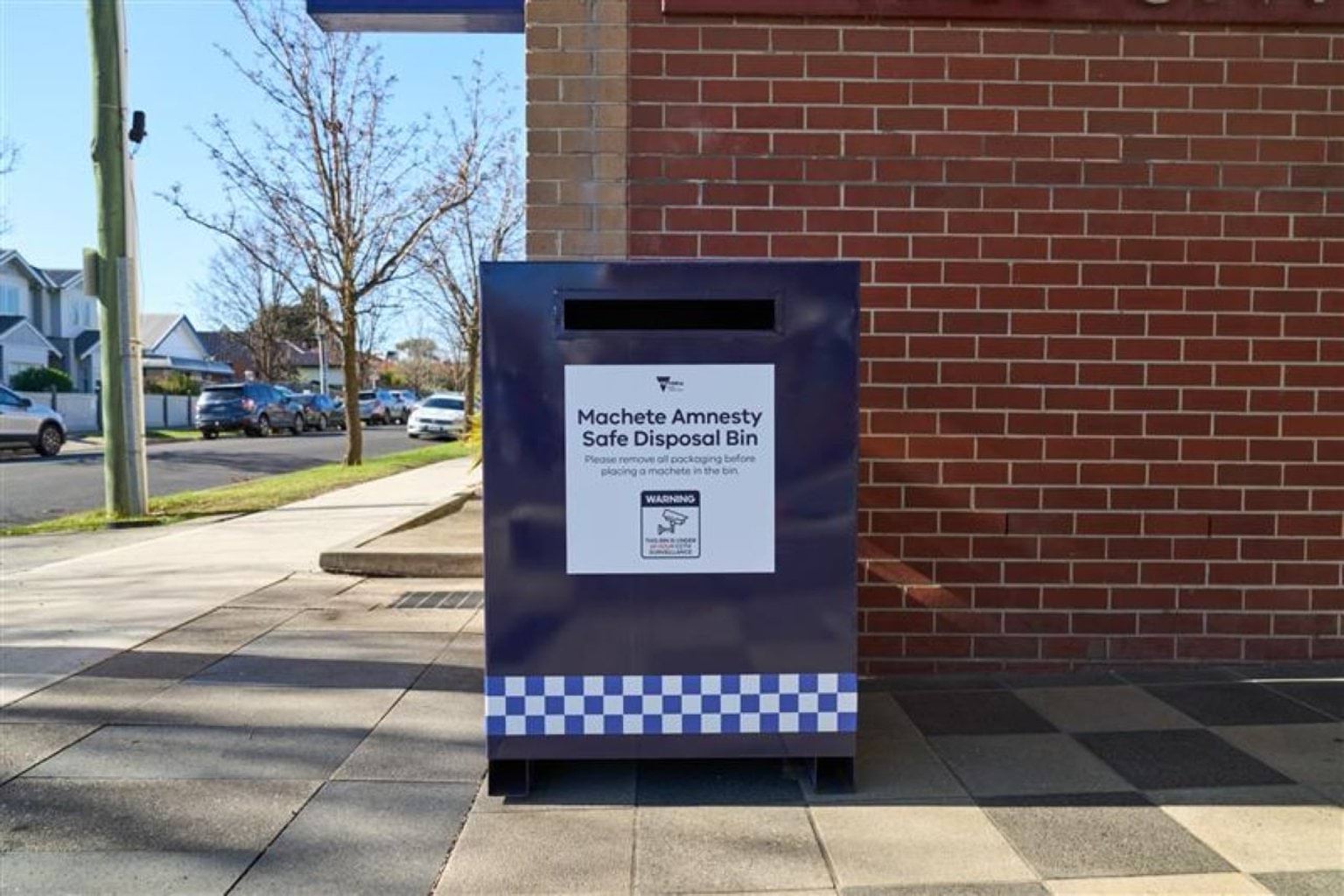 A large blue metal bin, labelled Machete Safe Disposal Bin, sits outside a Victoria Police station. There is a large slitted opening at the top of the bin for machetes to be dropped into. The sign asks to 'Please remove any packaging before placing a machete in the bin' and advises there are 24/7 cameras monitoring.