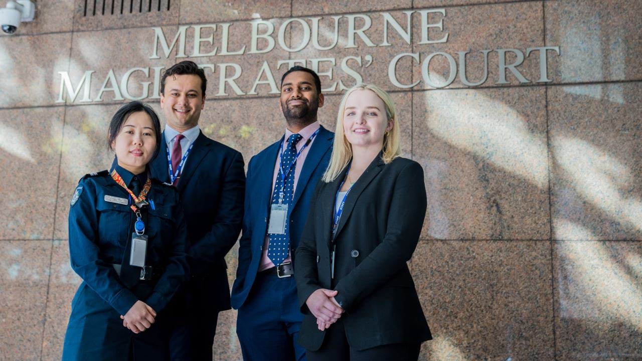 Three members wearing suits and one member wearing a Victoria Police uniform, stand in-front of the Melbourne Magistrates' Court. 