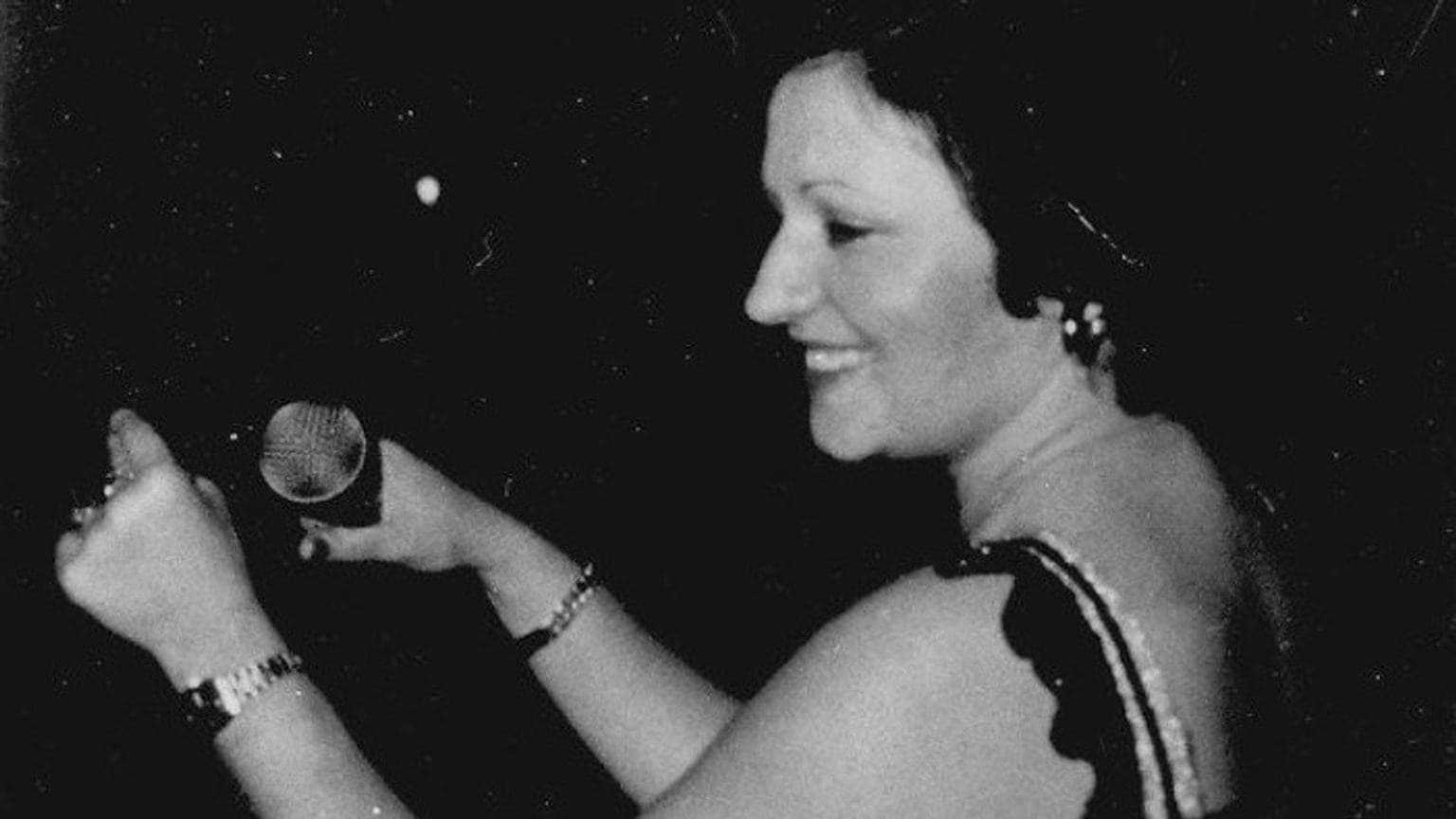 Haroula Kipouradou wearing a black strap dress, singing into a microphone 