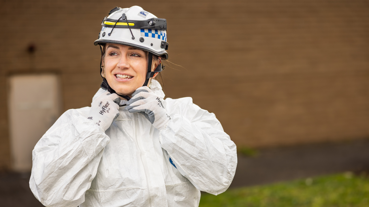 A female police officer dressed in a white forensic jumpsuit adjusts her safety helmet.