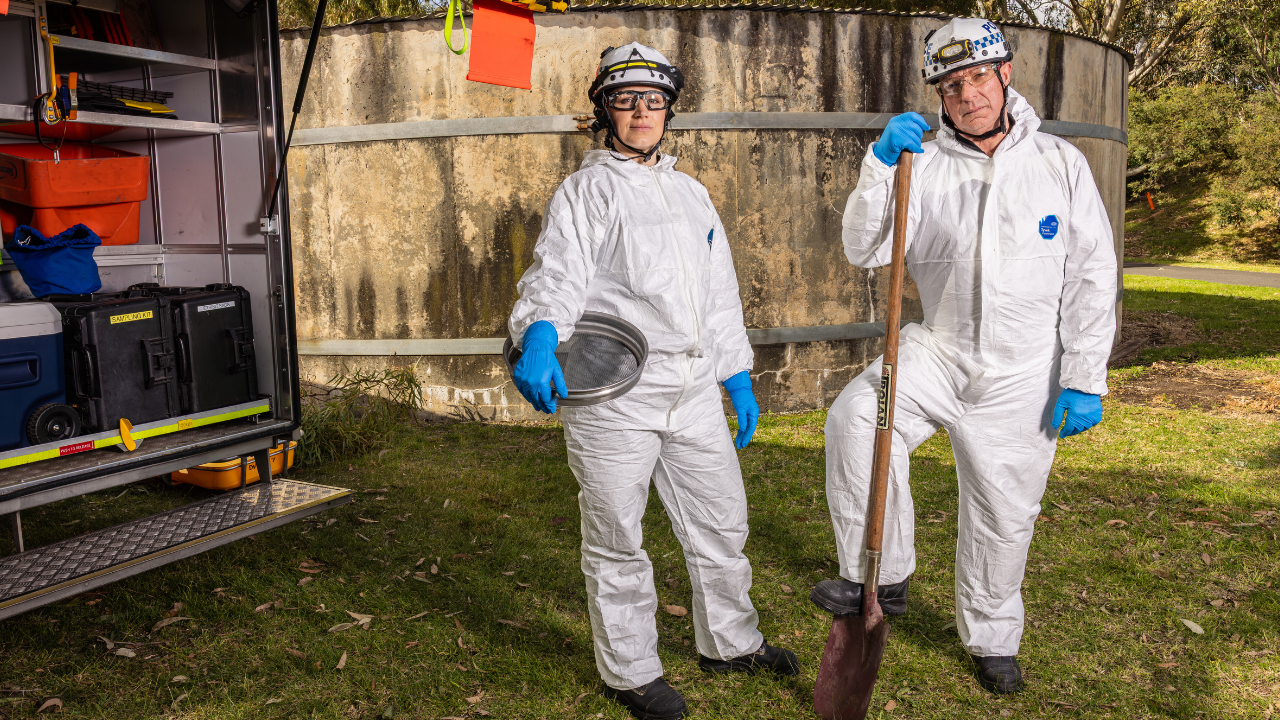 Two police officers dressed in white forensic jumpsuits, helmets and goggles smiling at the camera.