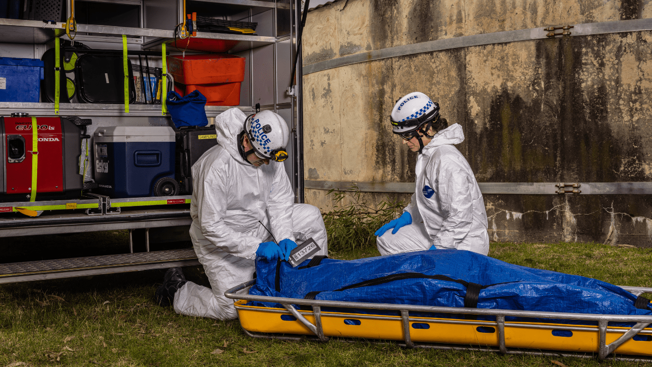 Two police members in white forensic jumpsuits and helmets kneel alongside a stretcher during a training scenario