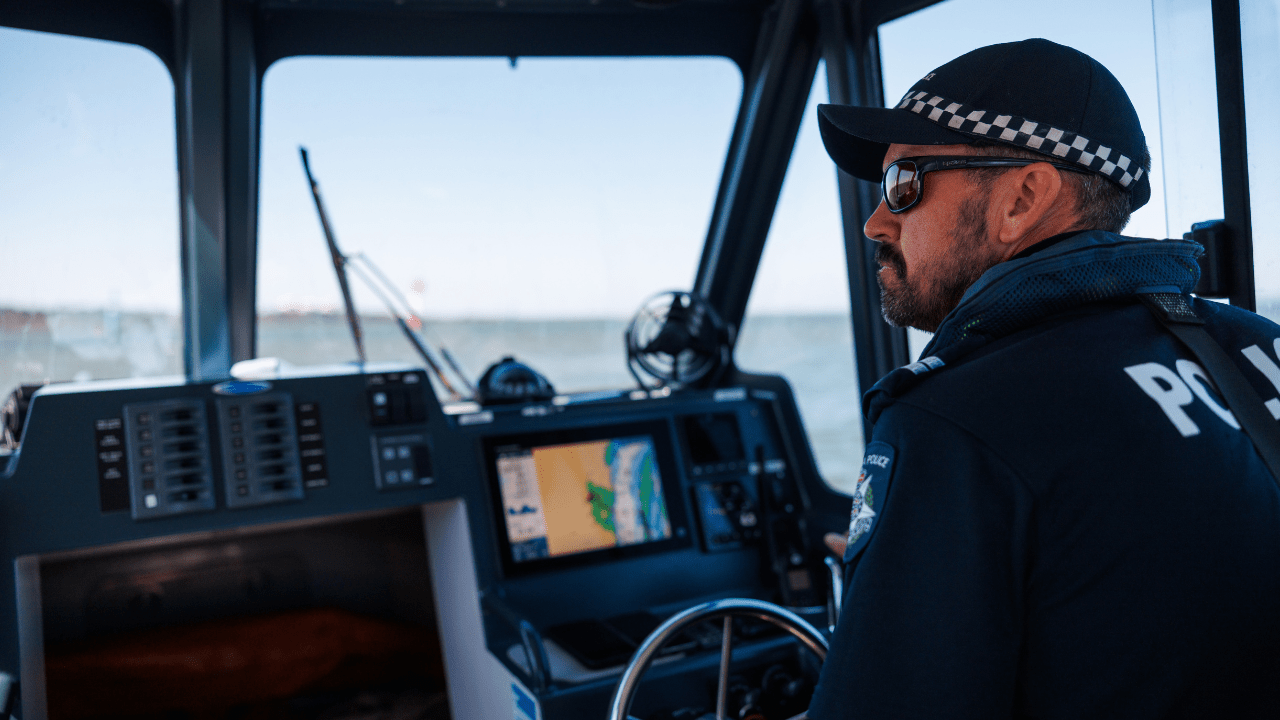 A male police officer in uniform wearing a cap and sunglasses looks out the front window of a police boat on Westernport Bay