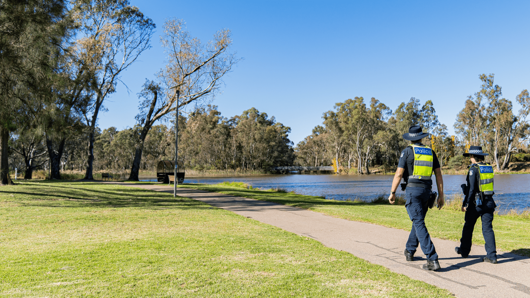 Two police officers in police uniform, walking in the sun on a path next to the river.