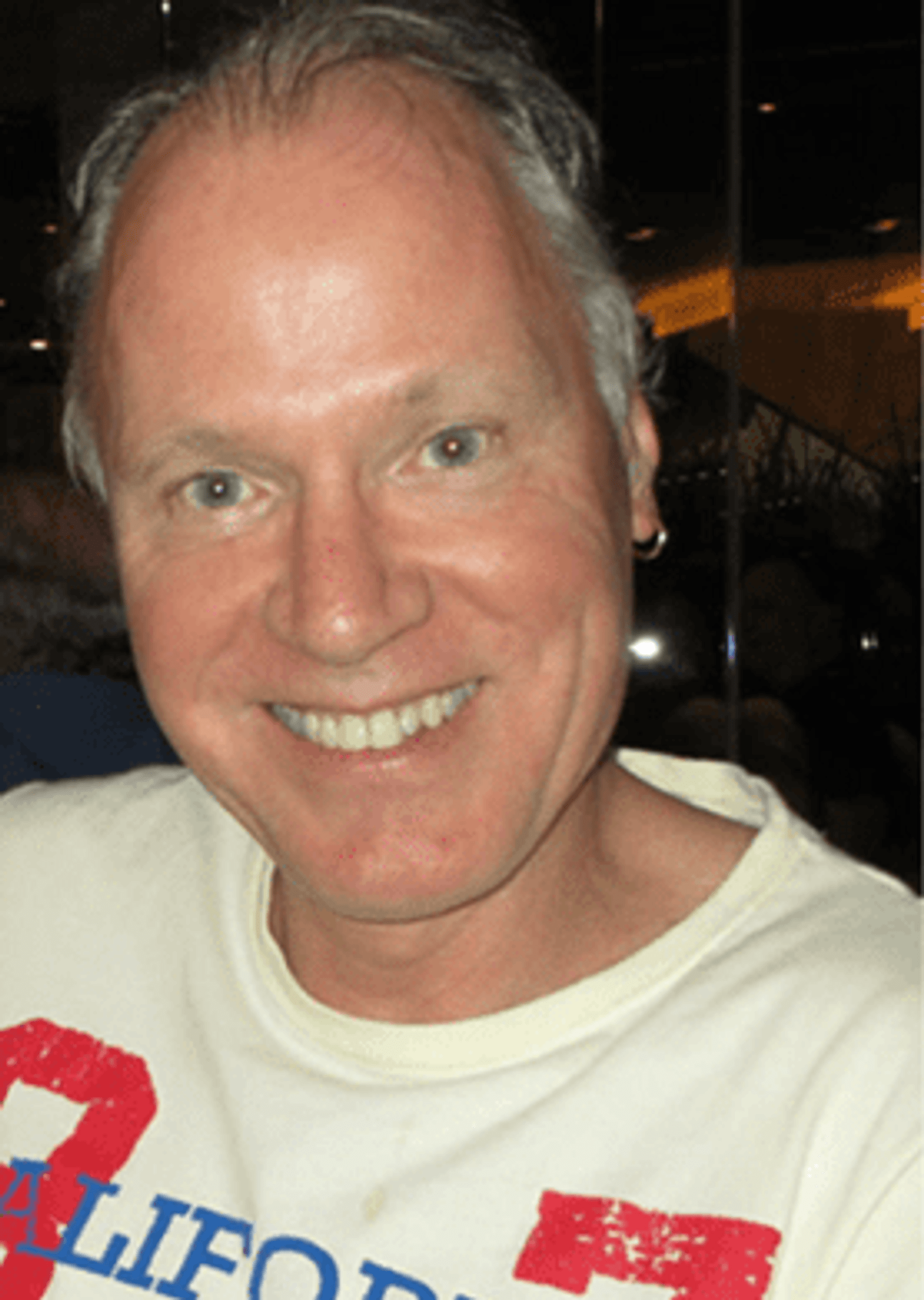 Brendan looking at the camera, with grey-blue eyes, thin grey/black hair and one hoop earring on his right ear, wearing a white t-shirt with California text on the front.