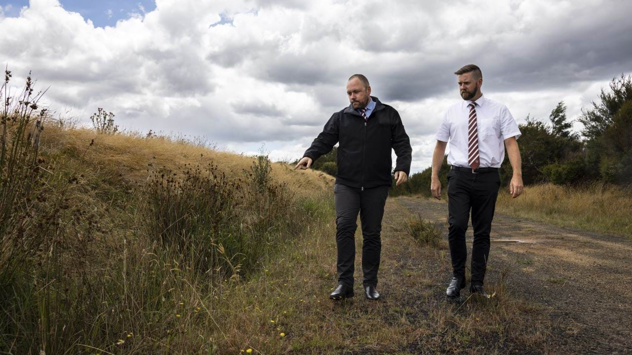 Two detectives in suits walk down a country road, where they believe missing person Robert Mather last travelled. . 