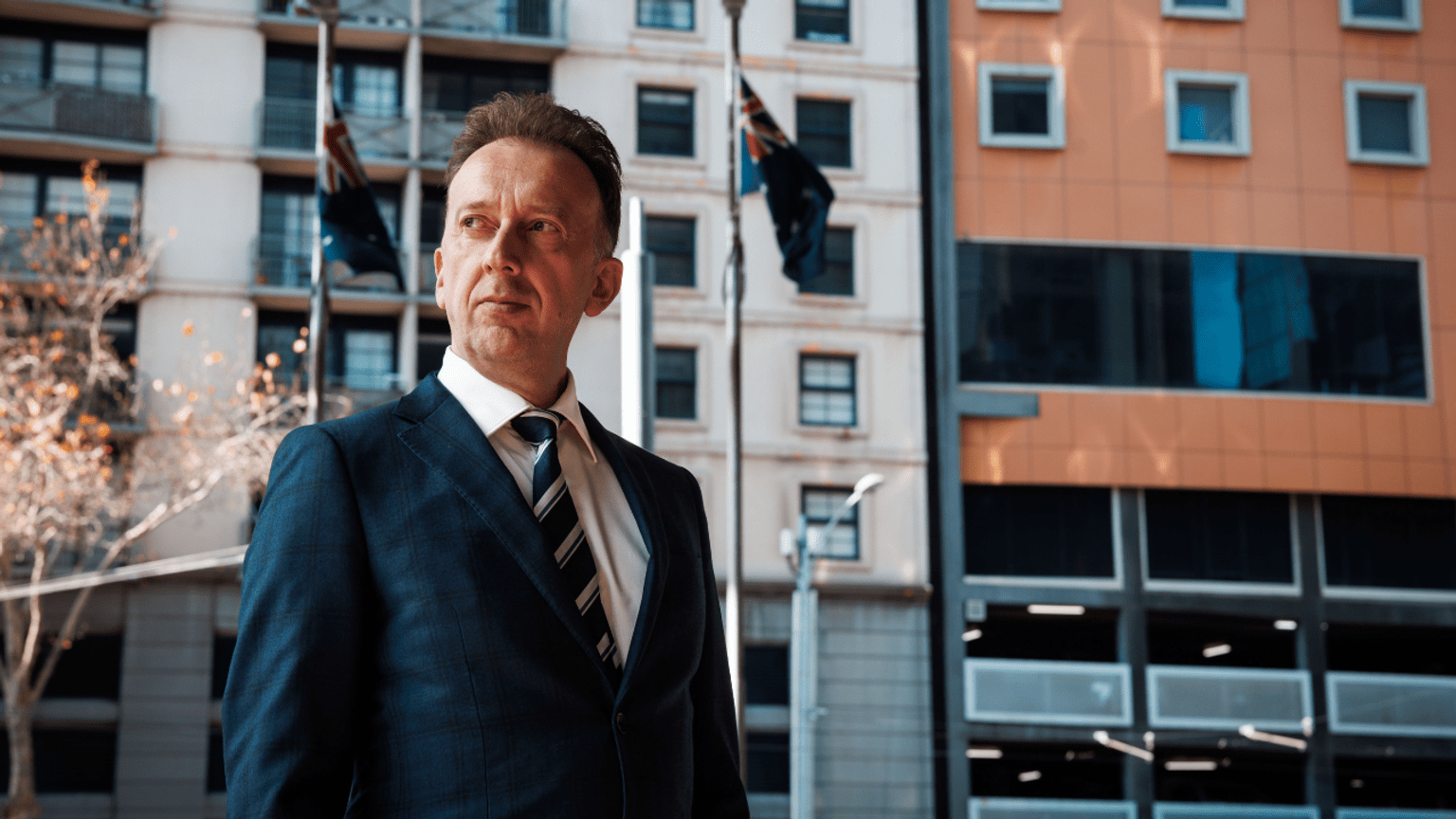 Profile image of Detective Sergeant Igor Rusmir, who is wearing a suit and tie and standing in front of flagpoles bearing the Australian and Victorian flags.