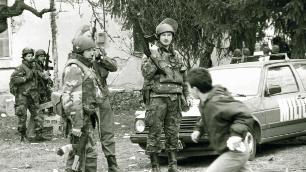 Men in camouflage holding weapons stand on a residential street in Pakrac in 1991 as a child runs past.