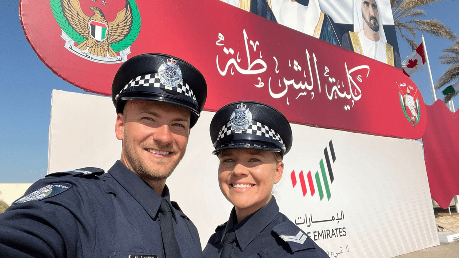 Police Life - Detective senior constables Mitch Lording and Megan Lewis, both dressed in Victoria Police uniform and hats, stand in front of a sign depicting Arabic wording in the United Arab Emirates.