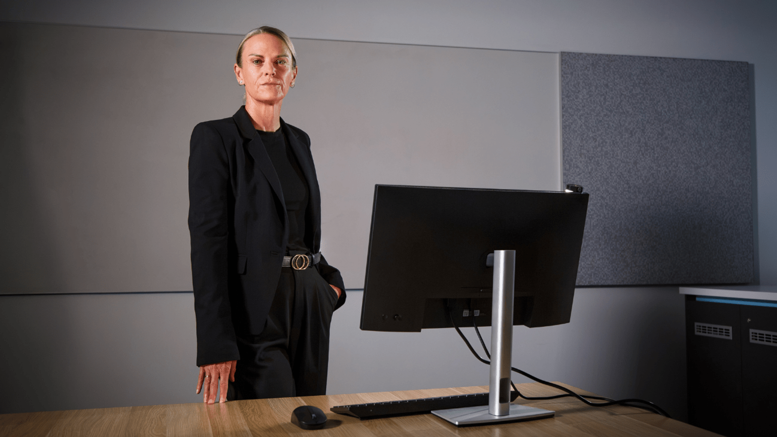 Det Ldg Sen Const Kellie Mervin, wearing a black suit, stands behind a desk with a computer monitor on it.