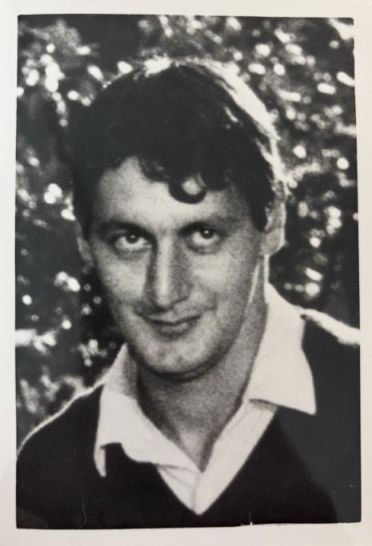 A black and white photo of Albert Pisani, wearing a collared shirt with a jumper over the top, he has short dark wavy hair.