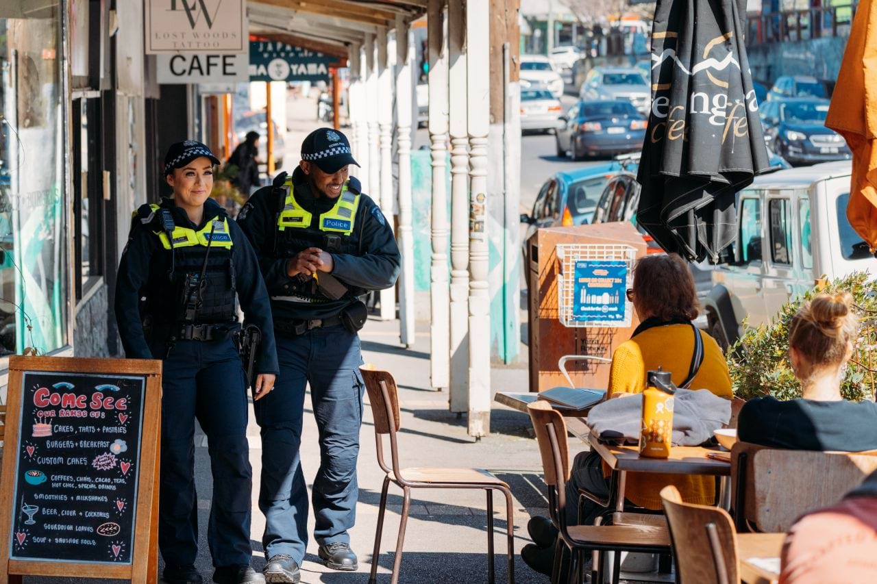 Two uniformed general duties officers smiling and walking down a suburban strip of shops, pausing to say hello to a community member seated at a nearby cafe. 