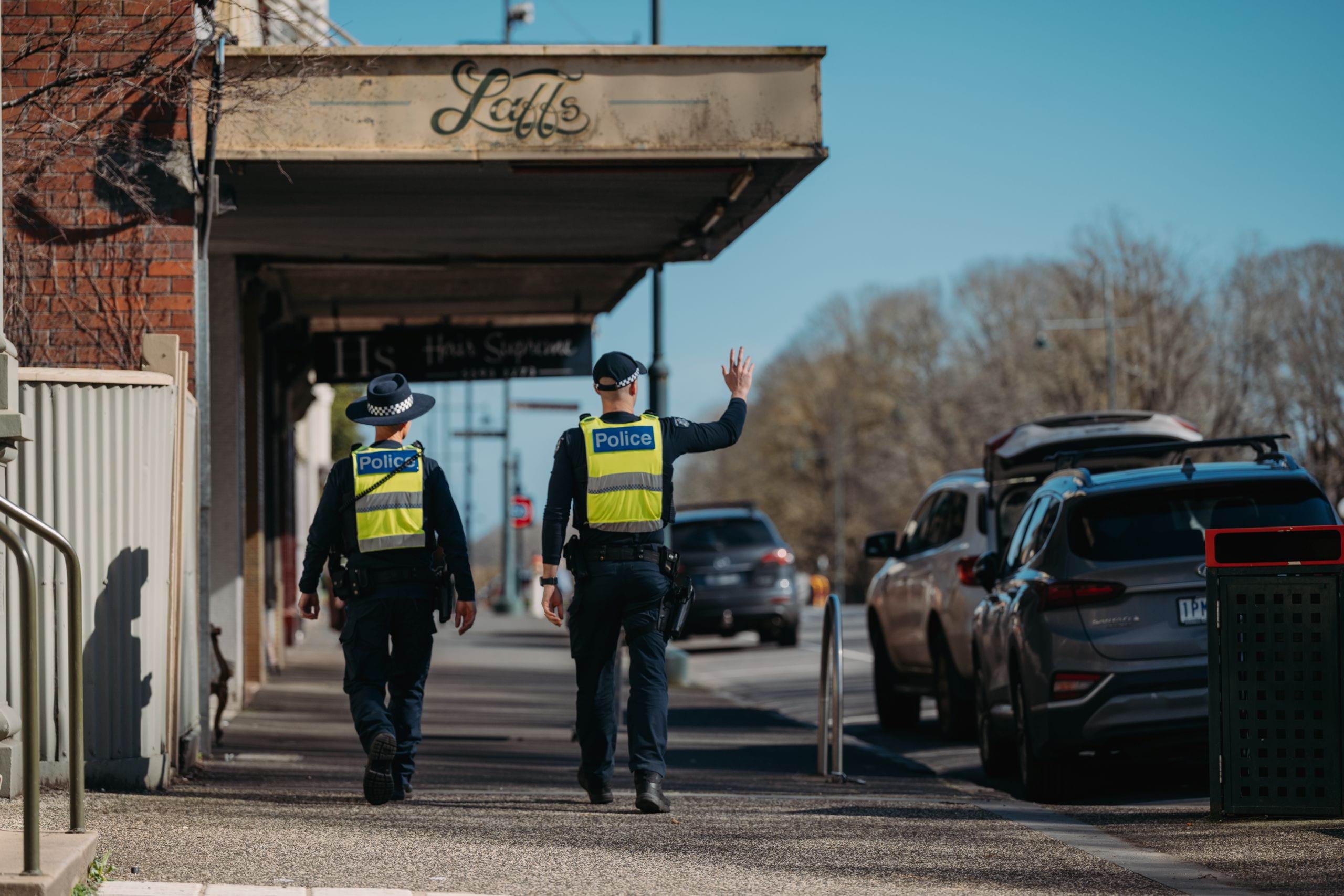 Two uniformed general duties officers walking away from the camera and down a sunny country town main street. One is wearing a wide brimmed hat, and the other is wearing a cap and waving to someone in the distance. 