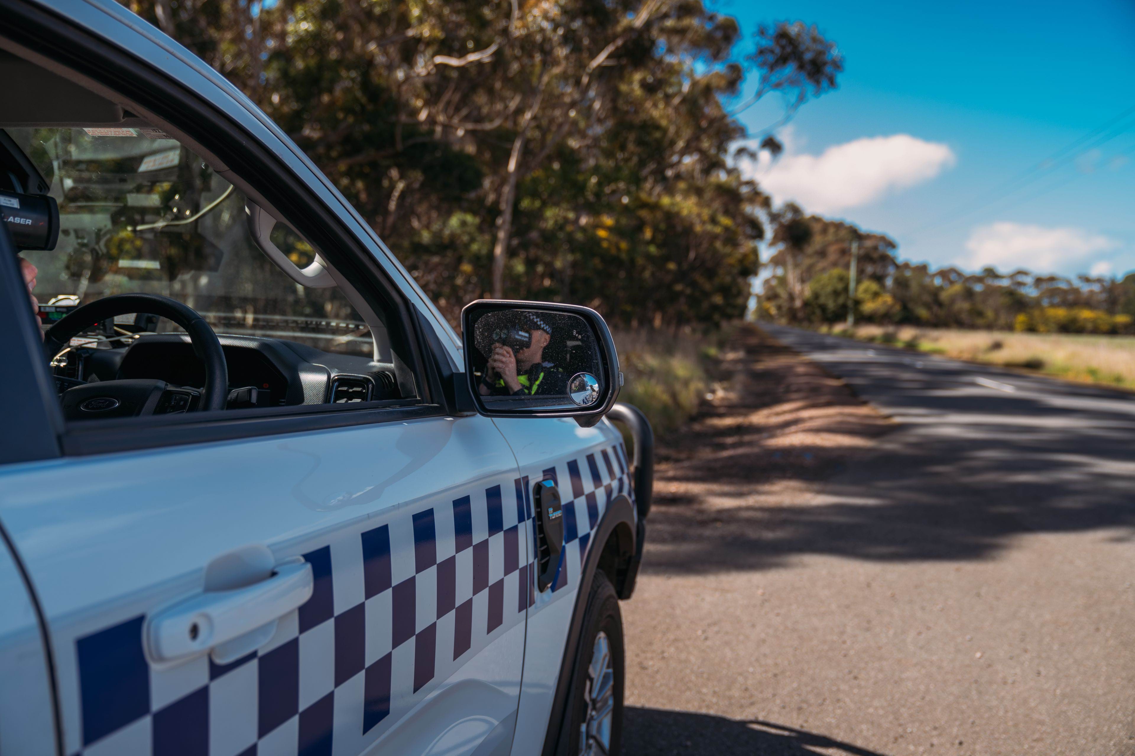 Parked police car facing the road, with a general duties officer holding a speed detection gun reflected in the side mirror. The car seems to be parked in a rural area with bush on one side and blue sky on the other. 