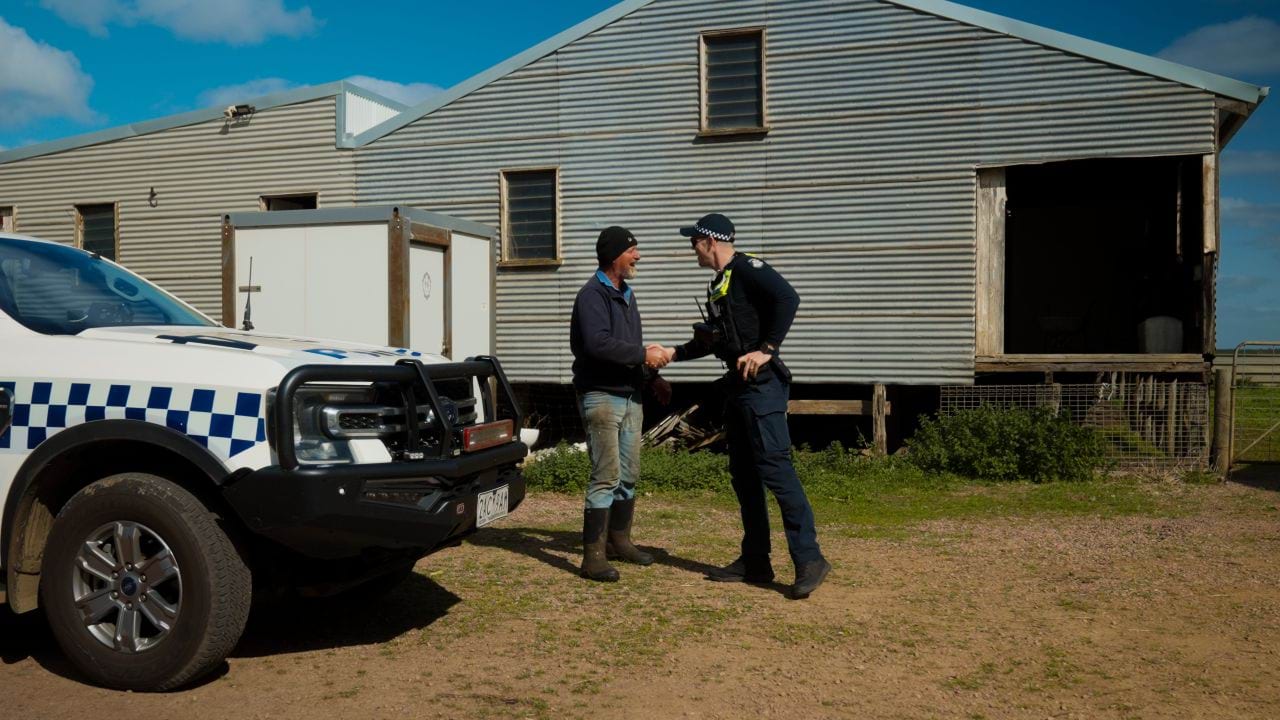 Farmer and uniformed police officer shaking hands and smiling on a farm, near a parked police car. There is a large shed in the background and sunny, blue skies. 